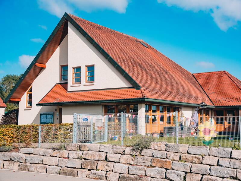 Die Ortsverwaltung und der Kindergarten in Bussmannshausen mit rotem Ziegeldach steht hinter einem Zaun und einer niedrigen Steinmauer, umgeben von blühenden Pflanzen, unter einem klaren blauen Himmel.
