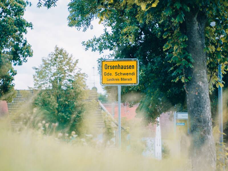 Ein gelbes Ortsschild mit der Aufschrift "Orsenhausen, Gde. Schwendi, Landkreis Biberach" steht zwischen Bäumen und Häusern.
