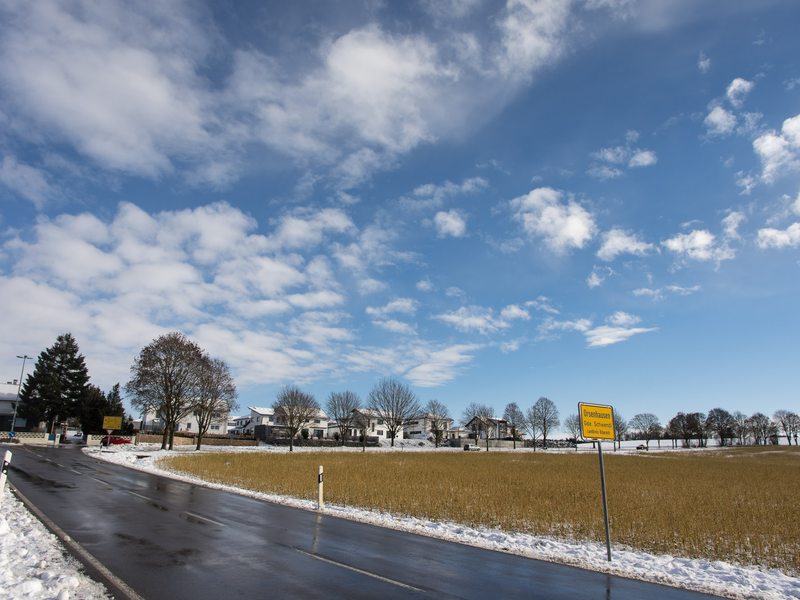 Eine verschneite Straße führt an einem Feld und dem Ortsschild von Orsenhausen vorbei, unter einem blauen Himmel mit vereinzelten Wolken.