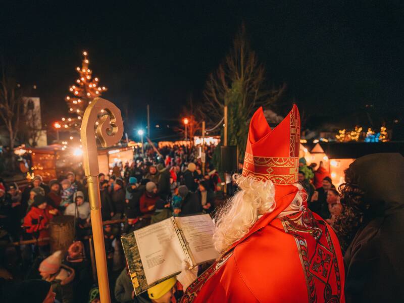 Der Hl. Nikolaus in einem roten Gewand und mit Mitra steht von hinten gesehen vor einer Menschenmenge auf einem weihnachtlich beleuchteten Markt.