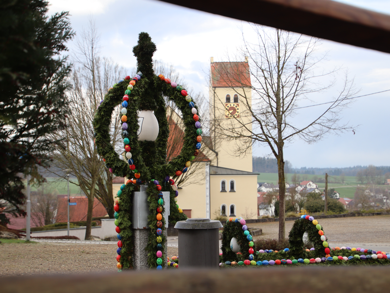 Mit Reißig und gefärbten Eiern geschmückter Brunnen. Im Hintergrund ist ein Kirchturm hinter Bäumen zu sehen