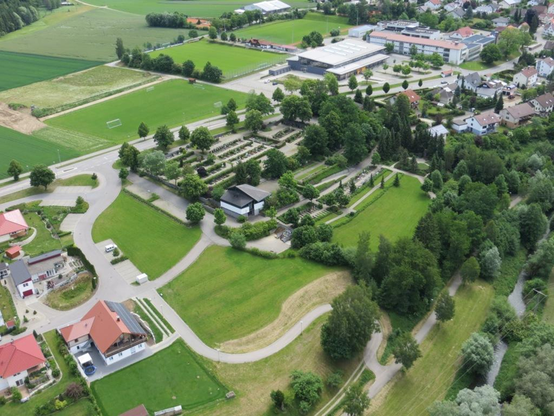 Luftbild im Vordergrund ist ein Teil des Wohngebiets zu sehen, in der MItte der Firedhof in Schwendi mit der Aussegungshalle. Im Hintergrund ist der Schulcampus in Schwendi zu sehen