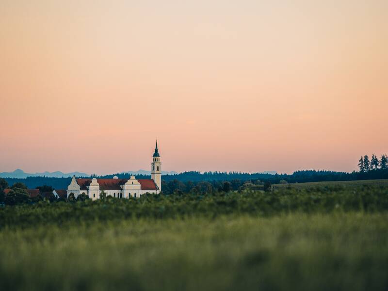 Eine weiße Kirche mit einem Turm steht in einer grünen Landschaft unter einem sanften, rosa gefärbten Himmel.