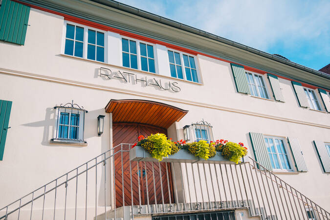 Das Bild zeigt die Fassade des Rathauses in Schwendi mit grünen Fensterläden und Blumenkästen vor dem Eingang.