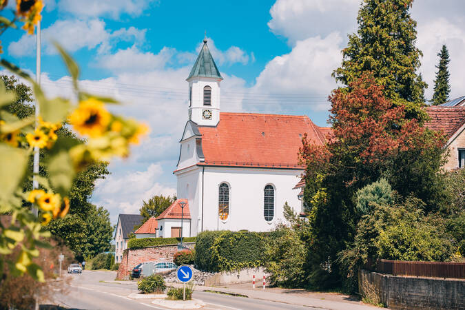 Eine malerische Kirche mit einem roten Dach und einem Turm erhebt sich auf einem von Pflanzen umgebenen Dorfweg, während im Vordergrund Sonnenblumen blühen.