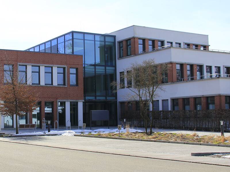 Ein großes Gebäude mit Wandelementen und Glaselementen an der Außenfassade. Davor eine Straße und Parkplätze. Links im Hintergrund folgt ein weiteres Gebäude.