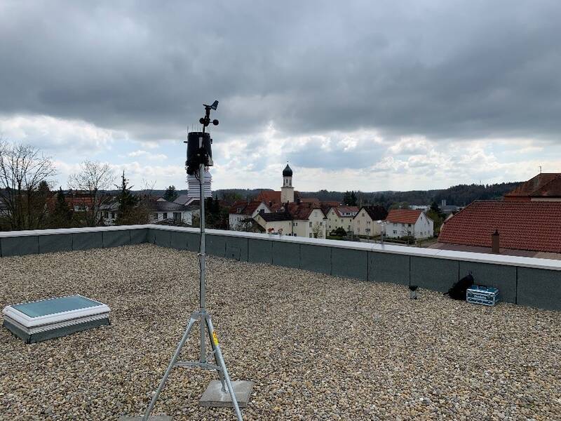 Auf einem flachen Dach, das mit Kieseln bedeckt ist., steht eine Wetterstation auf einem Stativ. Im Hintergrund sind Häuser und ein Kirchturm zu sehen.