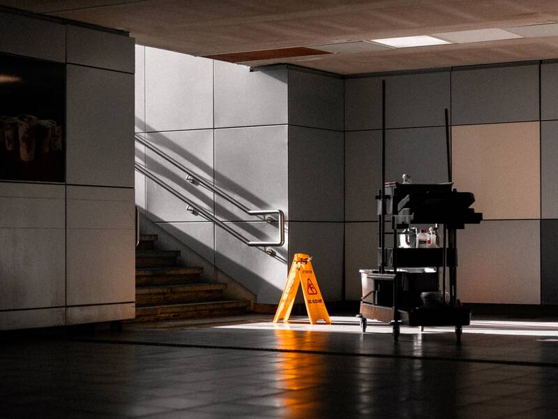 Ein gelbes Warnschild steht auf einem gefliesten Boden neben einer Treppe, die von Sonnenlicht beleuchtet wird.