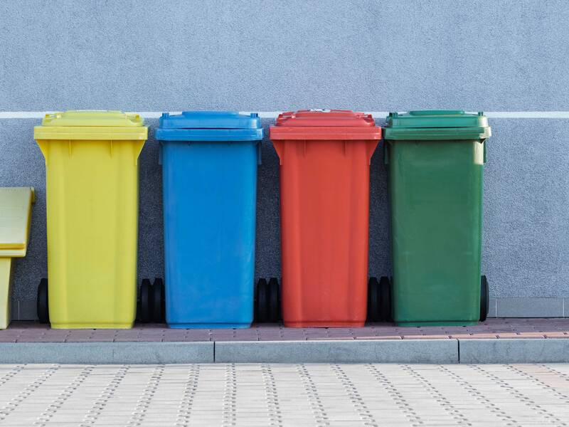 Vor einer grauen Wand stehend sind 4 Mülltonnen in gelb, blau, rot und grün zu sehen. Davor befindet sich ein gepflasterter Platz