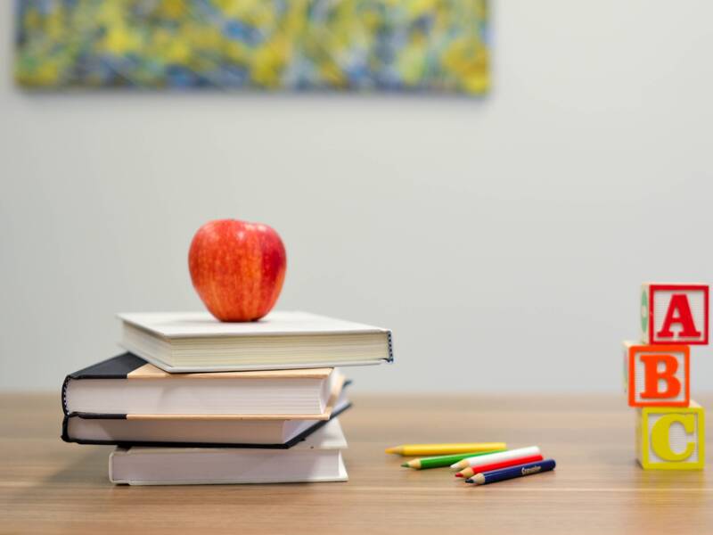 Auf einem Holztisch liegend auf der linken Seite ist ein Stabel von 4 Büchern zu sehen. Darauf liegt ein Apfel. Auf der rechten Seite ist ein Stapel aus drei Bauklötzen mit jeweils A,B und C. Zwischen den beiden Stapeln liegen mehrere Bundstifte.