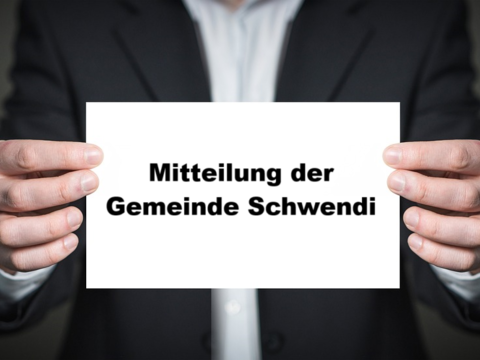 Ein Mann in Anzug dessen Kopf nicht zu sehen ist, hält mit beiden Händen ein weißes Blatt Papier in die Kamera. darauf steht: "Mitteilung der Gemeinde Schwendi"