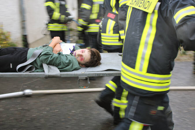 Auf einer Trage liegt ein Junge. Er ist als Verletzter geschminkt und hat die Augen geschlossen. getragen wird die Trage von Personen in einer schwarz gelben Feuerwehr Uniform. Auf deren Rücken ist Schwendi zu lesen. 