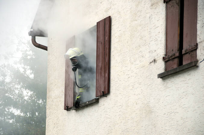 Feuerwehrmann unter schwerem Atemschutz neigt sich aus dem Fenster eines Hauses aus dem Rauch qualmt
