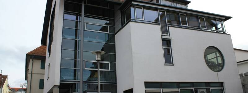 Bild des Rathauses. Blick vom Rathausplatz auf den Neubau. links ist eine große Glasfront zu sehen, die sich über alle drei Stockwerke zeiht. rechts davon ist eine grau-weiße Wand zu sehen. Unten und oben jeweils eine Glasfront und in der mitte ein großes Rundes Fenster. 