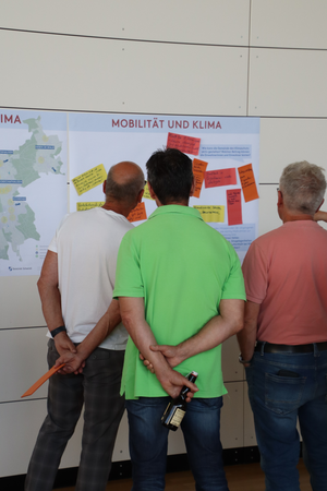 Drei Männer stehen vor einer Wand mit Notizen und Informationen zum Thema "Mobilität und Klima".