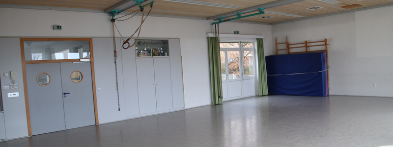 Bild aus dem Inneren des Gemeindesaals. Links ist die Eingangstür zu sehen, rechts davon Schränke in dem auch Pokale stehen. Rechts davon eine kleine Fenstereinheit und dann Sportmatten und eine Sprossenwand. Der Boden der davor zu sehen ist ist ein Hallenboden aus Linolium.