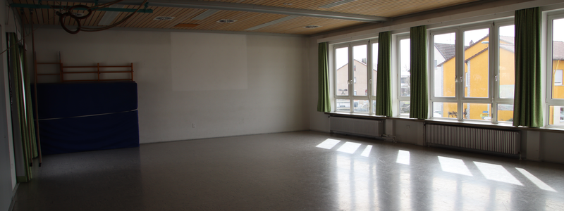 Bild aus dem Inneren des Gemeindesaals. links im Eck ist eine Sprossenwand zu sehen, vor der eine Dicke Matte steht. rechts ist eine große Fensterfront zu sehen. Der Boden ist ein typischer Hallenboden aus Linolium.