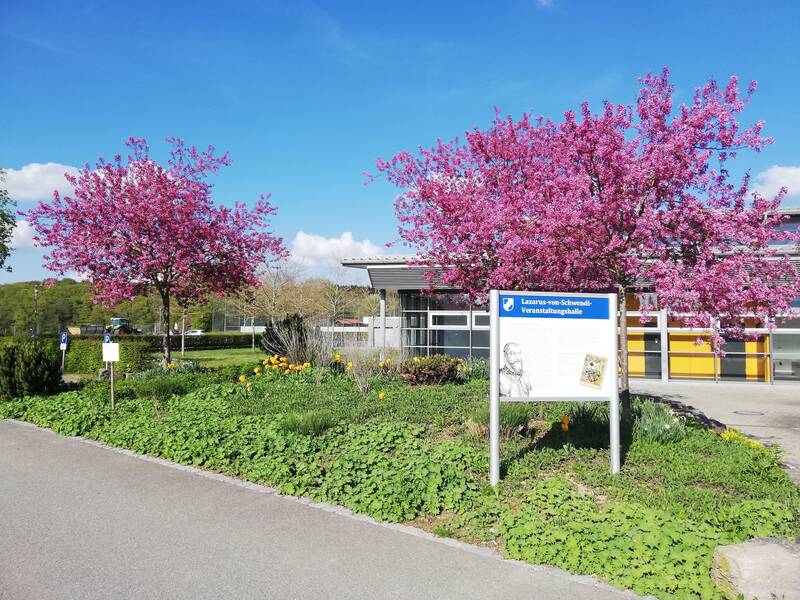 VHalle Schwendi mit Frühjahrsblütenbaum. In der Mitte des Bildes ist ein Grünes Blumenbeet zu sehen. Darin steht eine Informationstafel auf der Lazarus-von-Schwendi-Veranstaltungshalle zu lesen ist. Hinter der Tafel sind zwei ros blühende Bäume zu sehen. Hinter dem gesamten Beet ist ein Teil der Halle mit Flachem Dach zu erahnen.