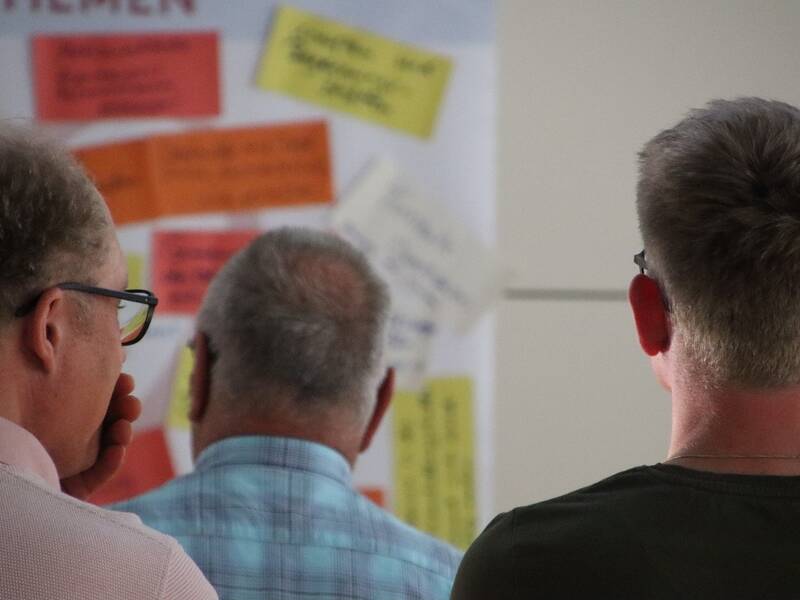 Drei Männer betrachten ein Whiteboard, das mit bunten Haftnotizen bedeckt ist.